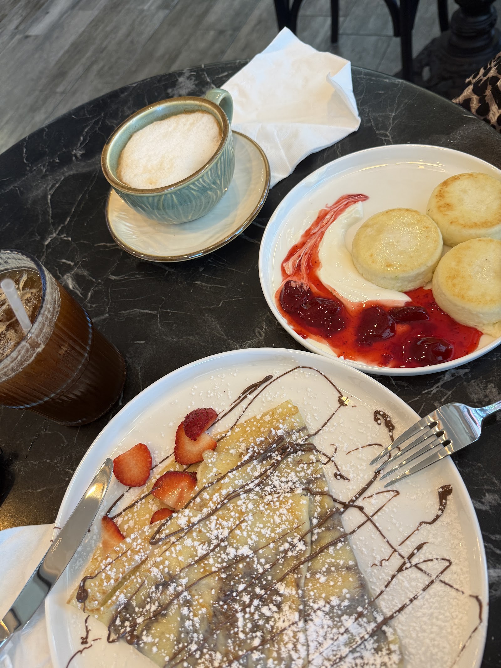 A table with a cappuccino, cheese pancakes with berry sauce and a crepe